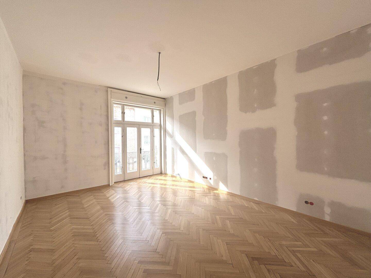 Helles Zimmer mit Fischgrätparkett und Renovierungsbedarf, viel Tageslicht durch große Fenster.