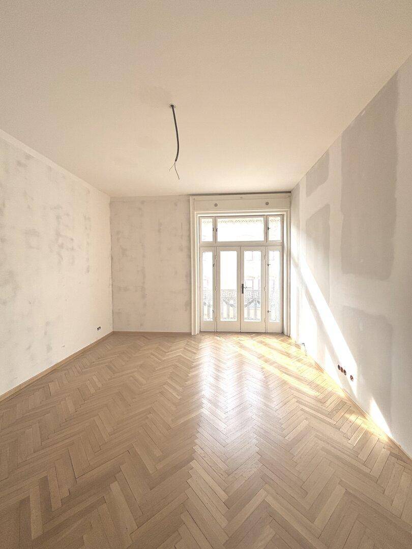 Lichtdurchflutetes Zimmer mit Fischgrätparkett und Renovierungsbedarf, Zugang zum Balkon.