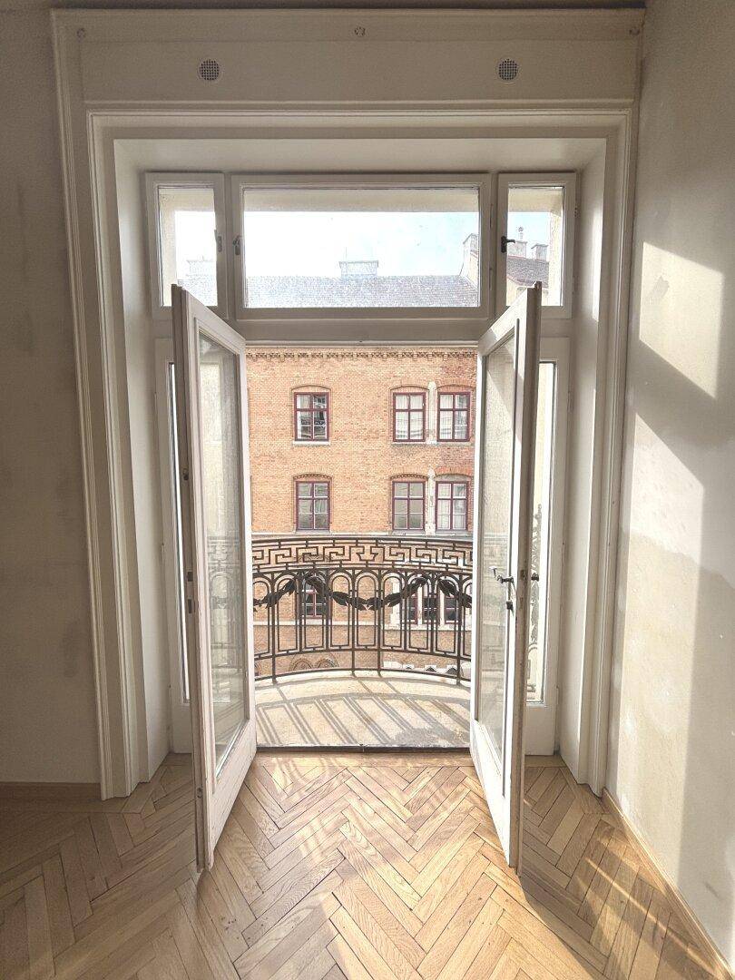 Helles Zimmer mit geöffneten Balkontüren und Blick auf die gegenüberliegenden Gebäude.