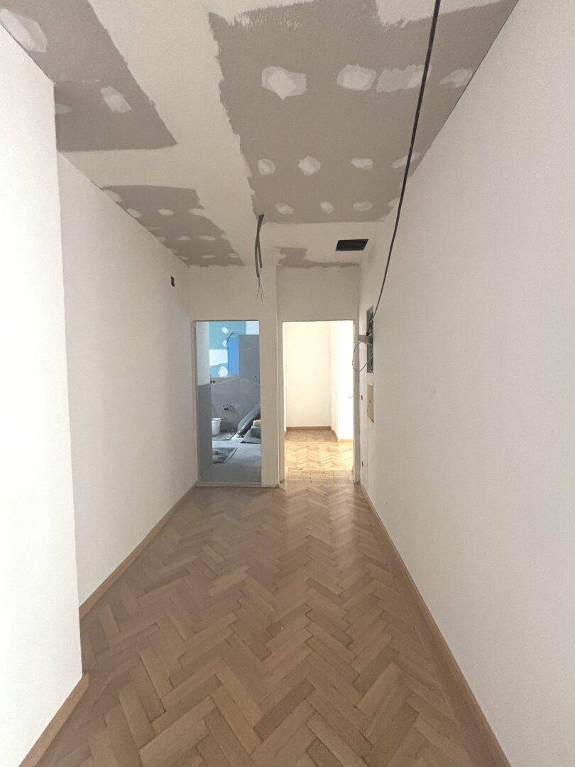 Langer Flur mit Fischgrätparkett und Renovierungsspuren, führt zu weiteren Räumen.