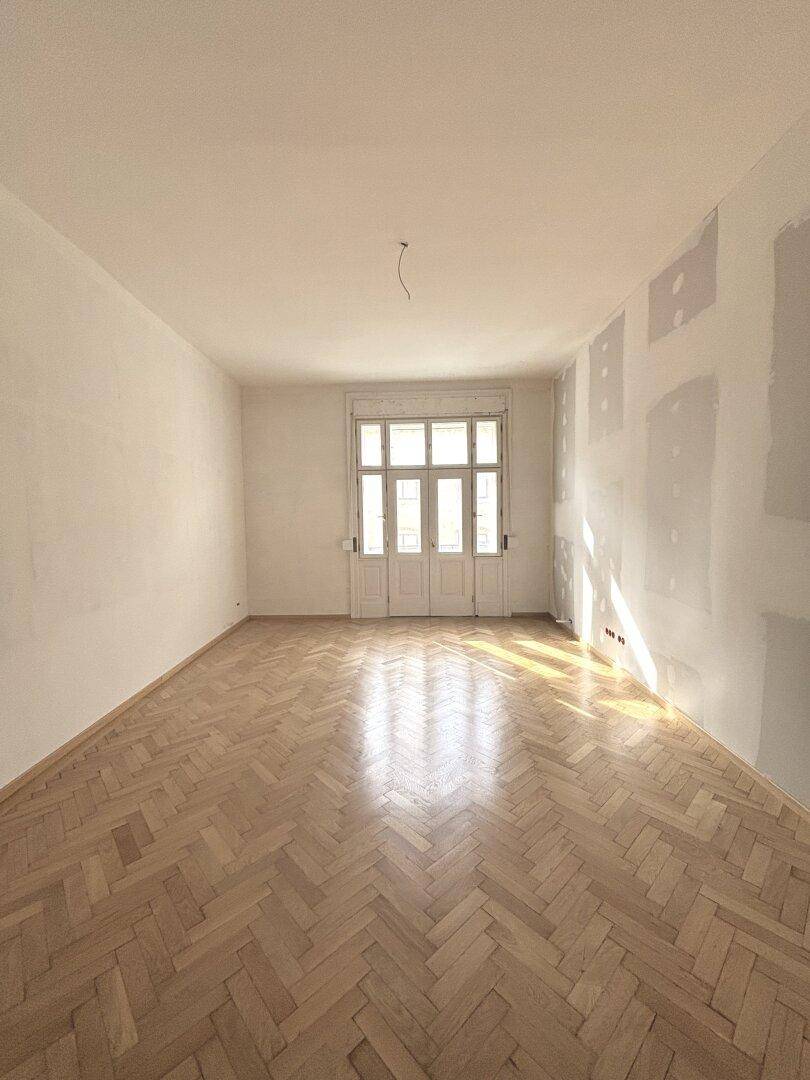 Großes Zimmer mit Fischgrätparkett und hellen Wänden, viel Sonnenlicht durch die Fenster.
