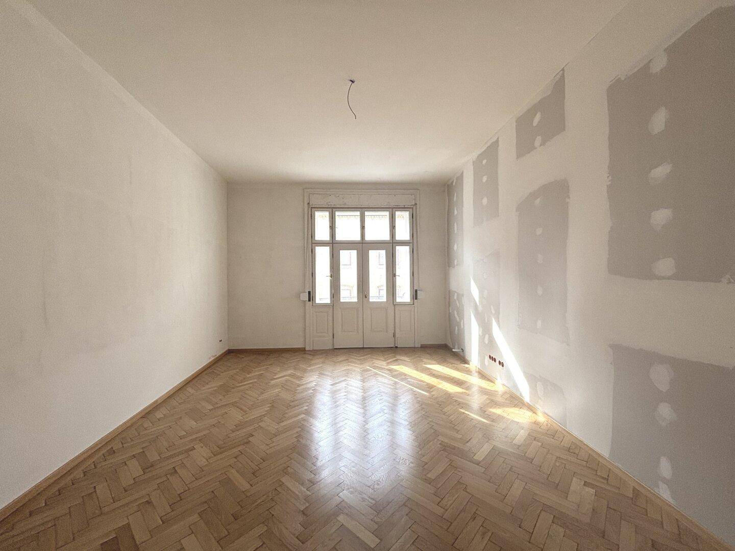 Geräumiges Zimmer mit Fischgrätparkett und Renovierungsspuren, heller Lichteinfall durch große Fenster.