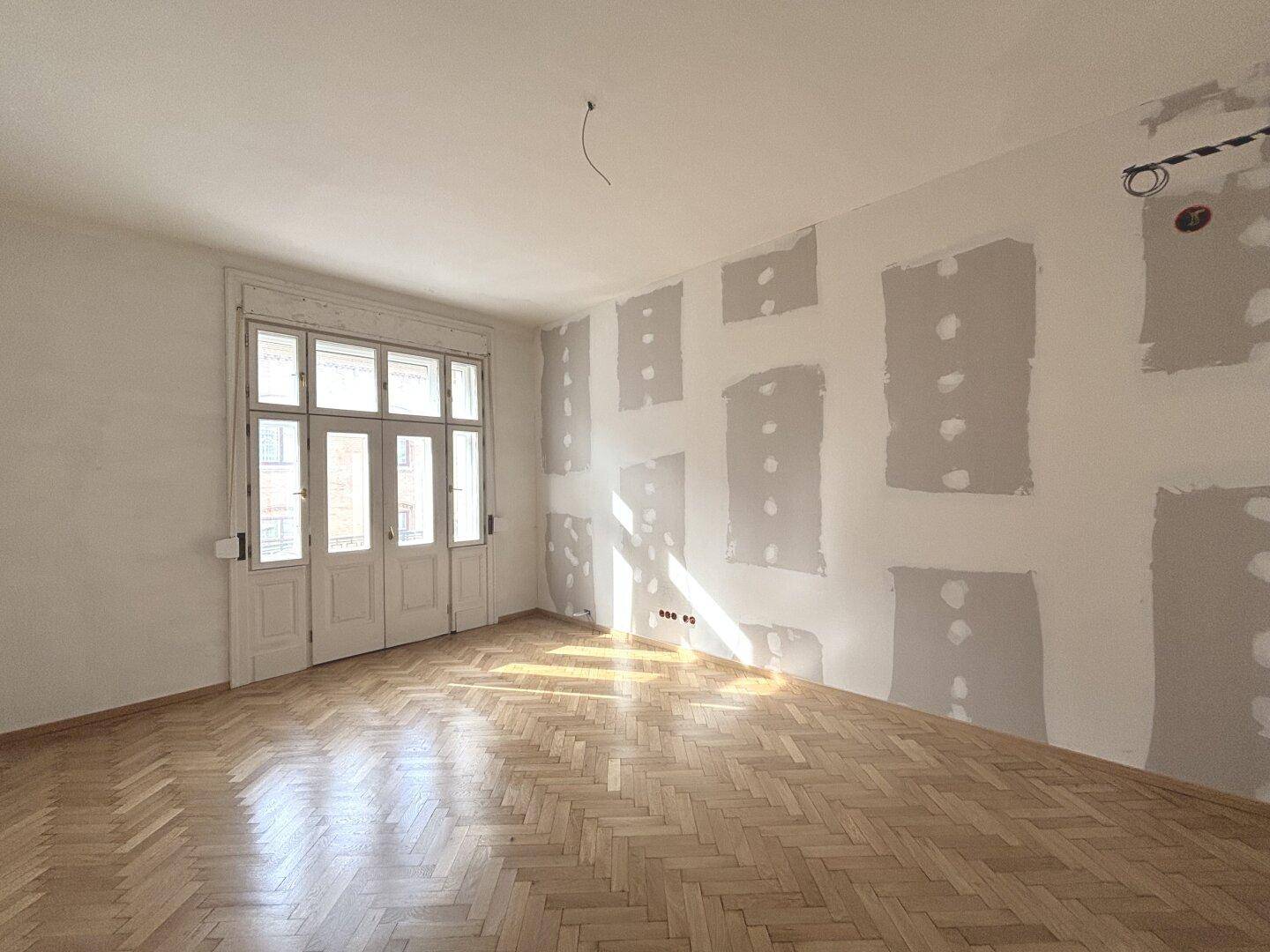 Geräumiges Zimmer mit Fischgrätparkett und Renovierungsbedarf, viel Tageslicht durch die Fenster.
