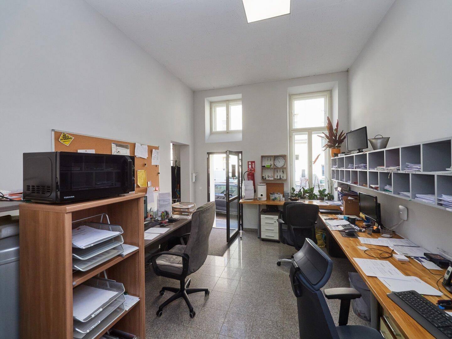 Heller Arbeitsbereich mit mehreren Schreibtischen, Bürostühlen und großen Fenstern für Tageslicht.