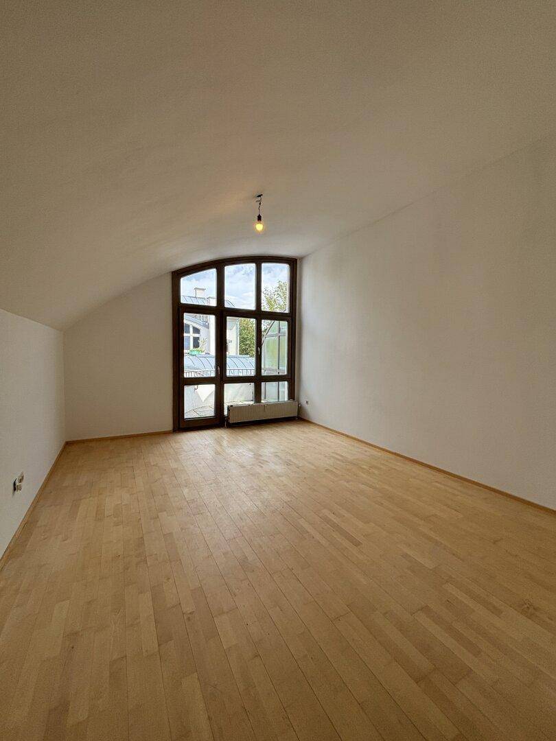 Geräumiges Zimmer mit hellem Holzfußboden, Dachschräge und einem großen Fenster für viel natürliches Licht.