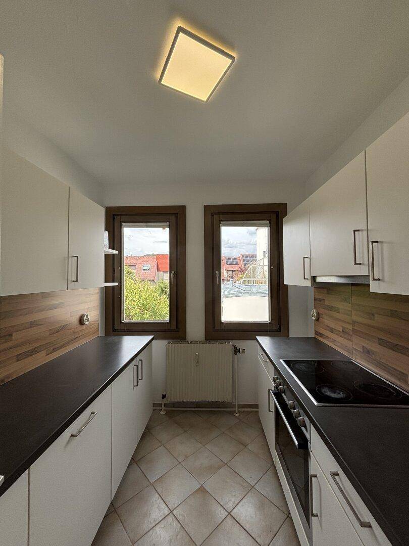 Funktionale Küche mit weißen Schränken, dunklen Arbeitsplatten und Fliesenboden, hell durch zwei Fenster.