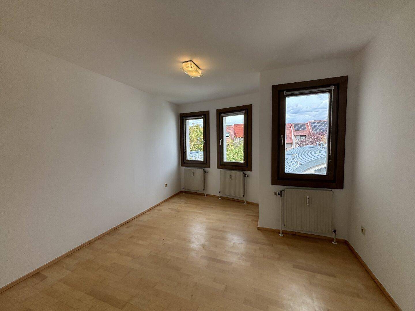 Helles Schlafzimmer mit Holzfußboden und drei Fenstern, die einen Blick nach draußen ermöglichen.