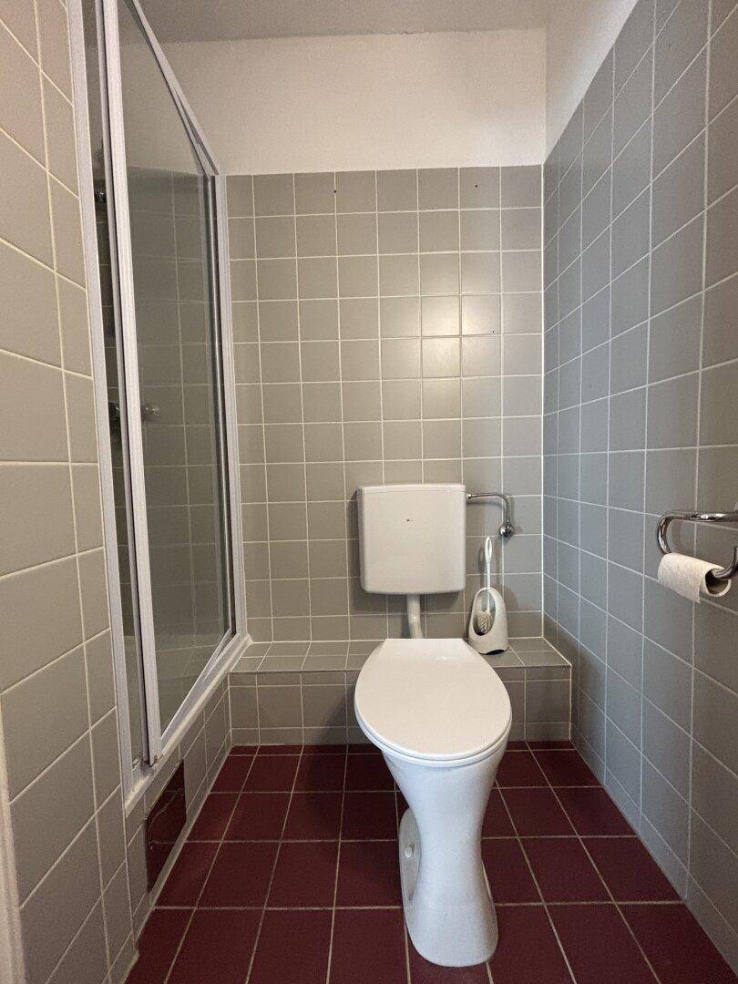 Kleines Badezimmer mit WC und Duschkabine, ausgestattet mit grauen Wand- und roten Bodenfliesen.