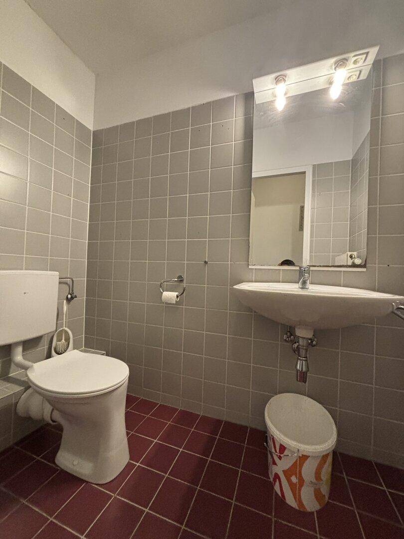 Kleines Badezimmer mit WC, Waschbecken und Spiegel, gestaltet mit grauen Wand- und roten Bodenfliesen.