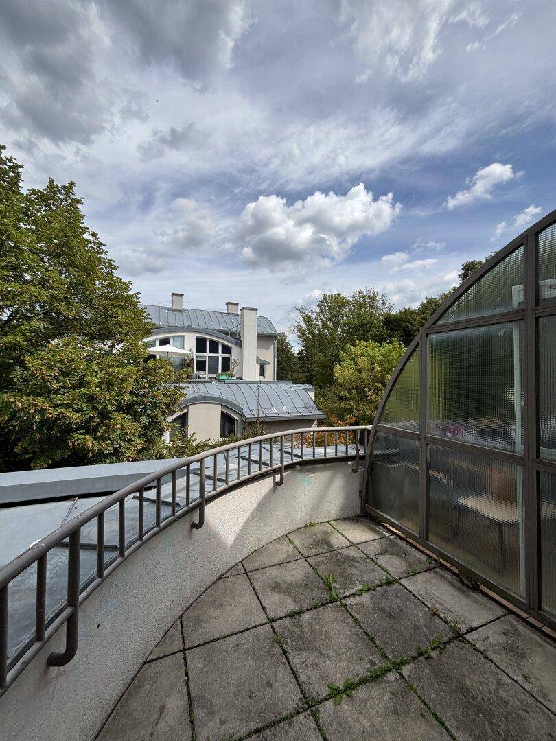 Terrasse mit Fliesenboden und Glasdach, bietet einen Ausblick auf die grüne Umgebung und Gebäude.