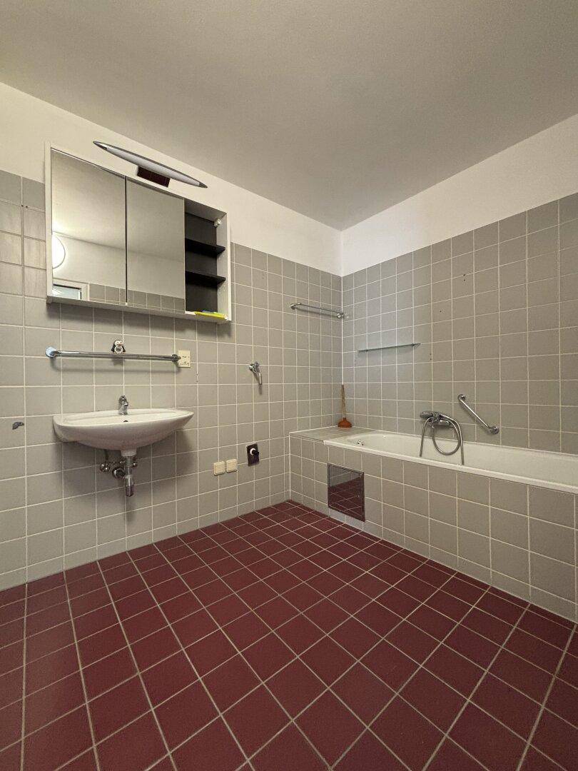 Badezimmer mit grauen Wandfliesen und roten Bodenfliesen, ausgestattet mit Badewanne und Waschbecken.