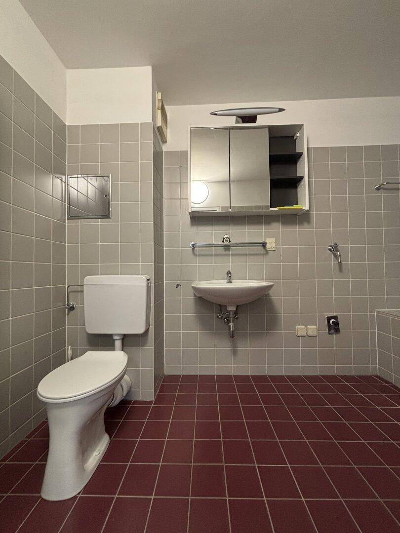 Badezimmer mit WC, Waschbecken und Badewanne, charakterisiert durch graue Wand- und rote Bodenfliesen.