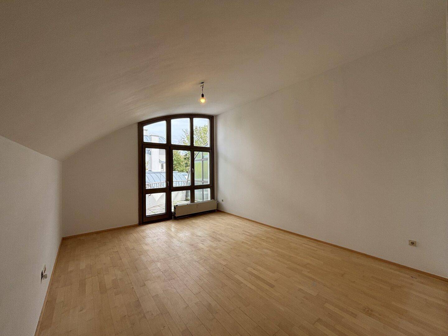Geräumiges Zimmer mit hellem Holzfußboden, Dachschräge und einem großen Fenster, das viel Licht spendet.