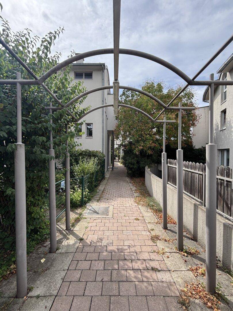 Gepflasterter Weg mit Metallbögen, umgeben von Grünflächen und führt zwischen Gebäuden hindurch.