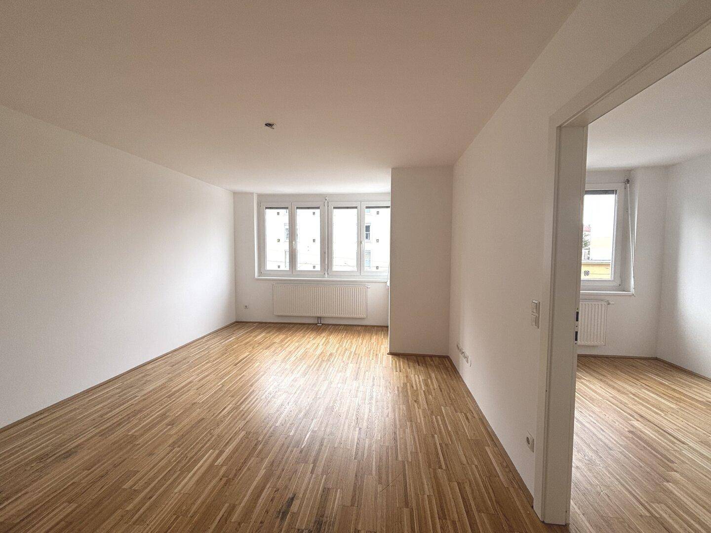 Leerer Wohnbereich mit Parkettboden und Fenstern, die viel Tageslicht hereinlassen.