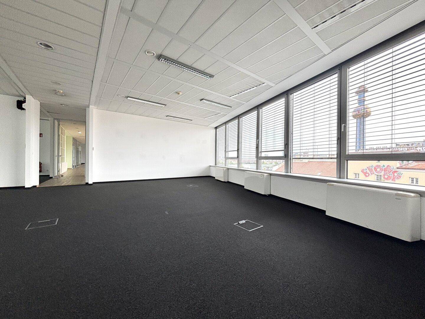 Geräumiger Büroraum mit Teppichboden, großen Fenstern und Stadtblick.
