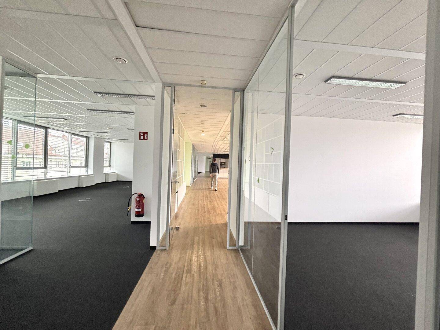 Bürogang mit Holzboden und Glastrennwänden, der zu weiteren Büroräumen führt.