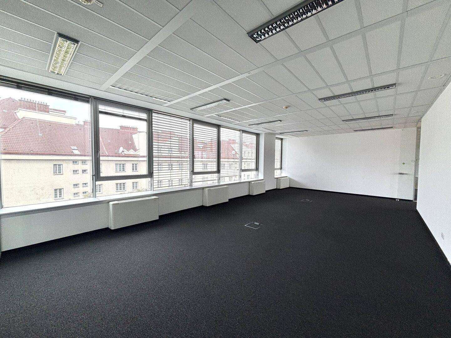 Heller Büroraum mit Teppichboden, großen Fenstern und Blick auf die umliegenden Gebäude.