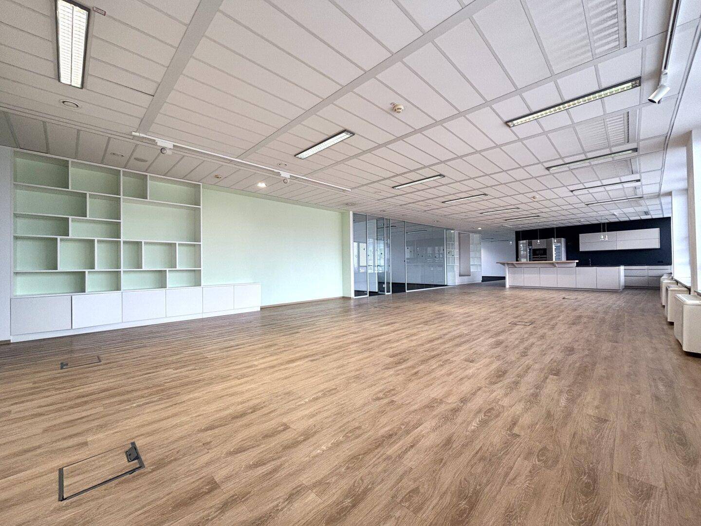 Offener Bürobereich mit Holzboden, integrierten Regalen und Blick auf eine moderne Küchenzeile.