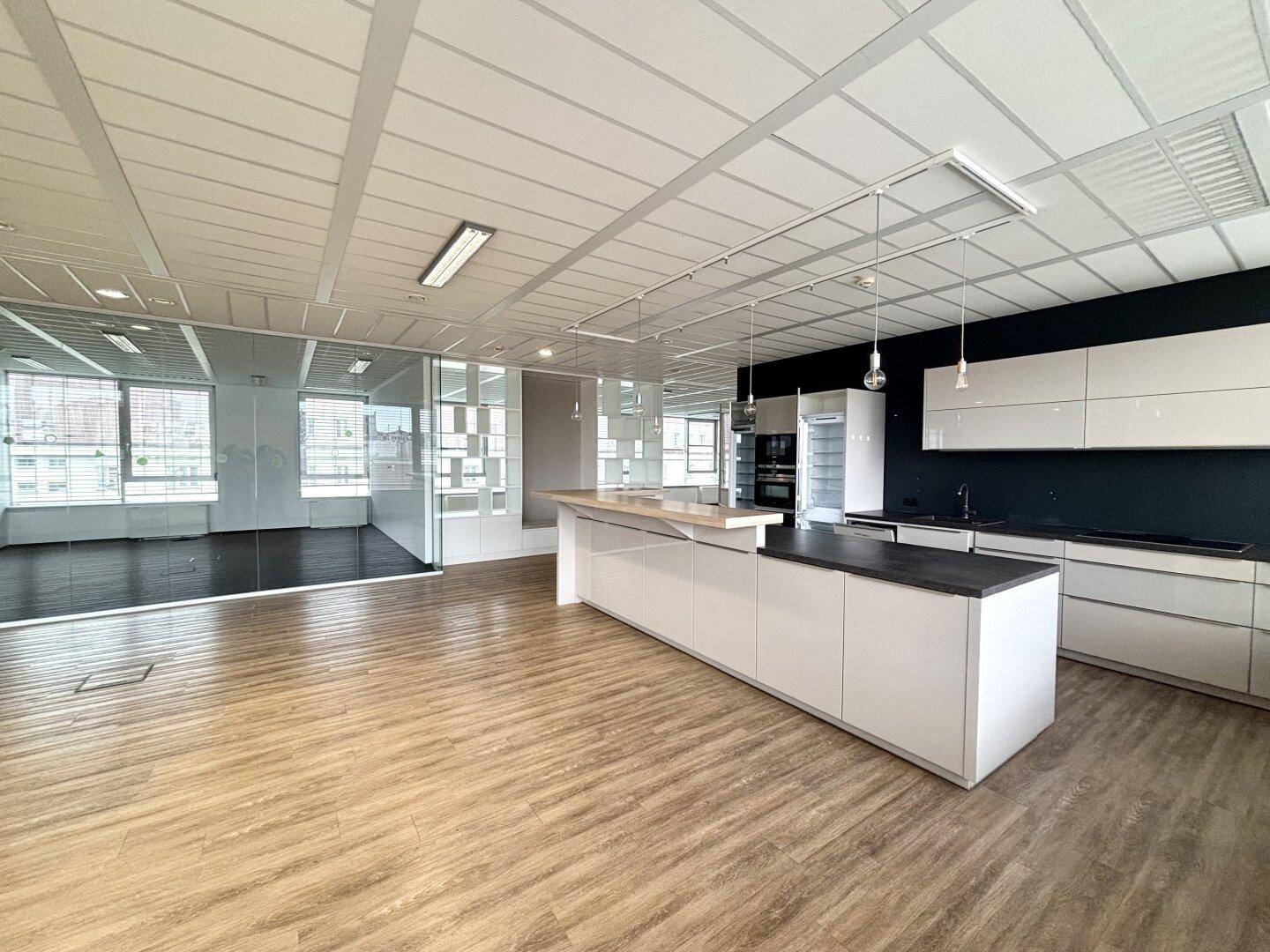 Moderne Küchenzeile in einem offenen Bürobereich mit weißen Schränken und dunkler Arbeitsplatte.