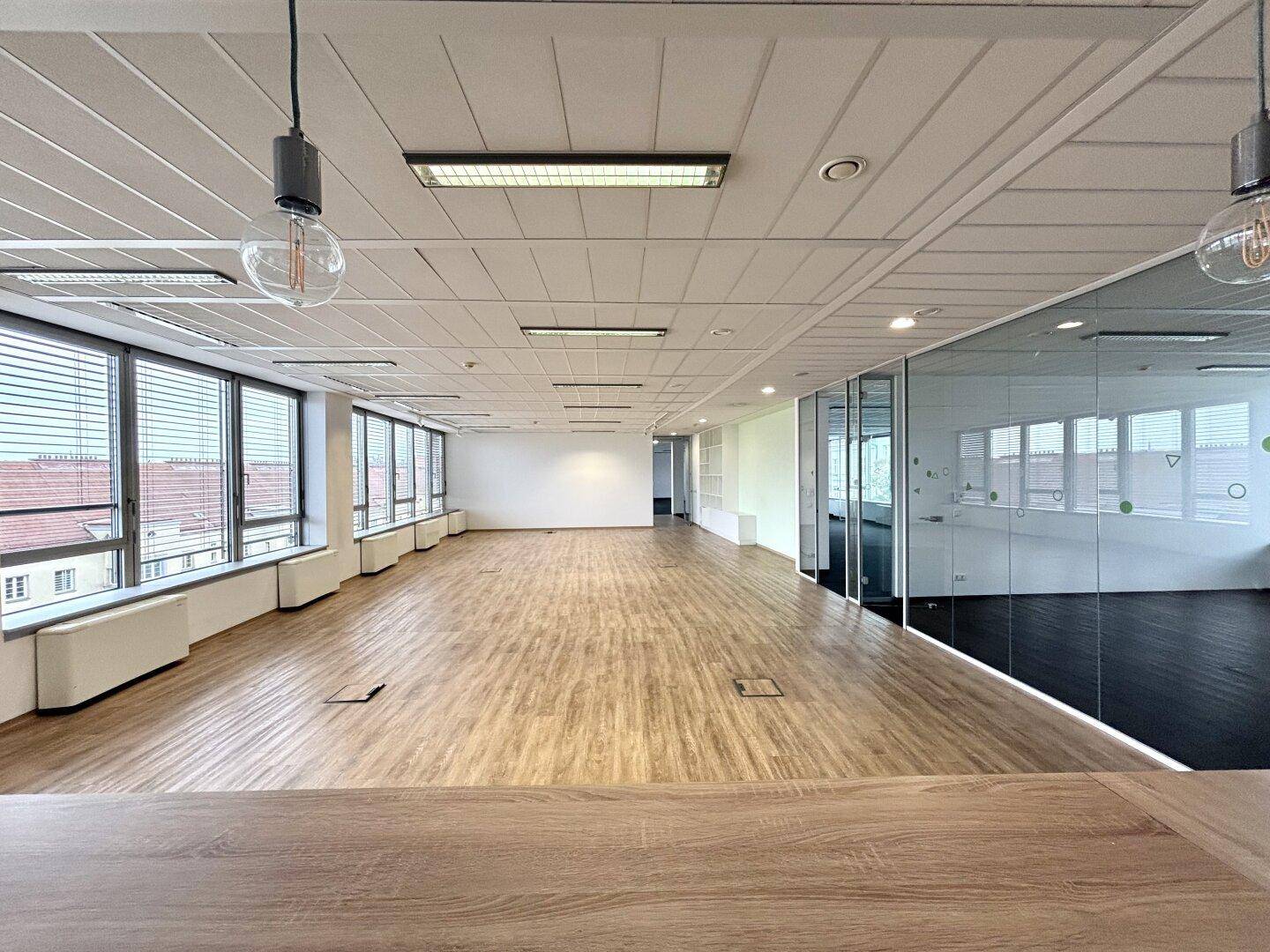 Heller, offener Bürobereich mit großen Fenstern, Holzboden und abgehängter Decke.