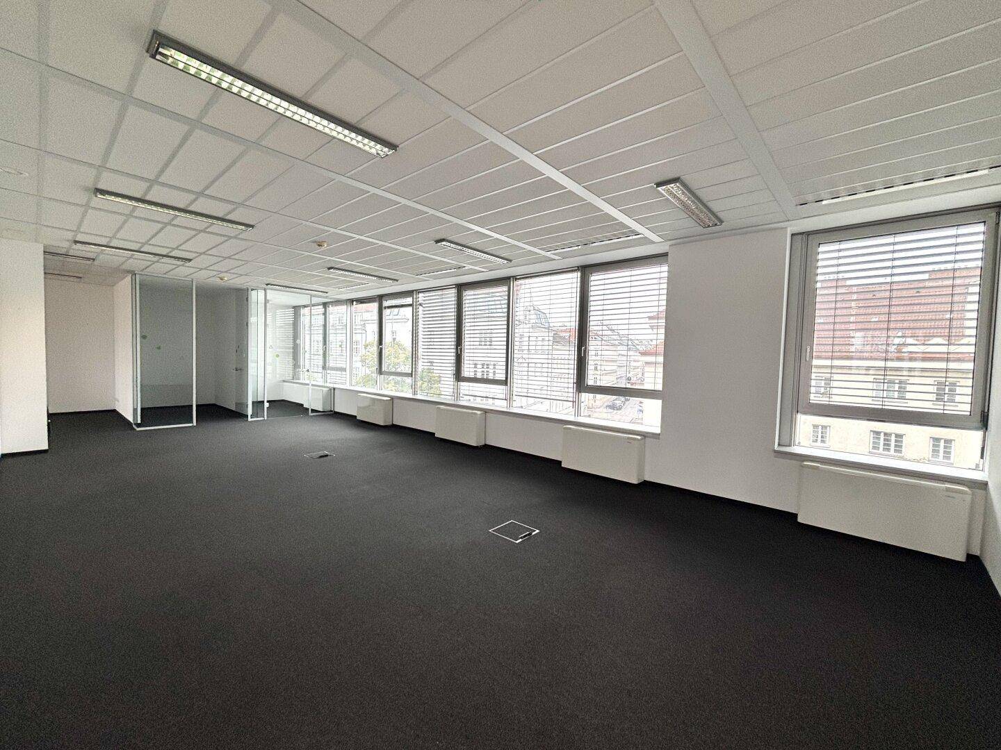Geräumiges, leeres Büro mit Teppichboden, Rasterdecke und großen Fenstern mit Jalousien.