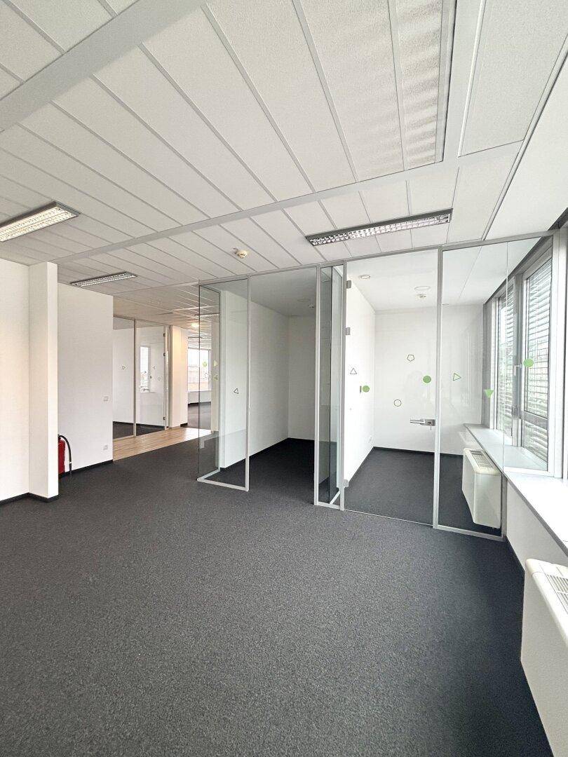 Heller Büroraum mit Teppichboden, Glasabtrennungen und Blick auf weitere Bürobereiche.