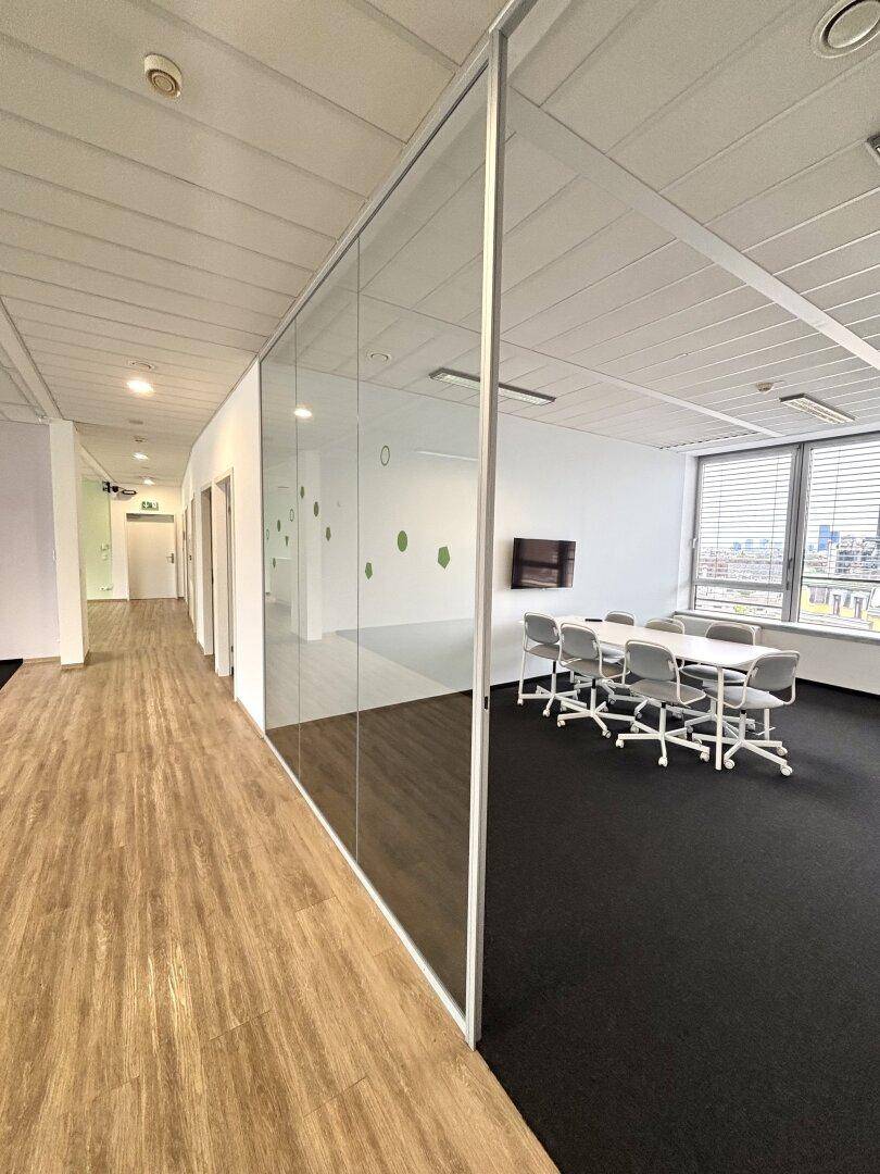 Konferenzraum mit Glastrennwand, Teppichboden und Blick auf den langen Flur.