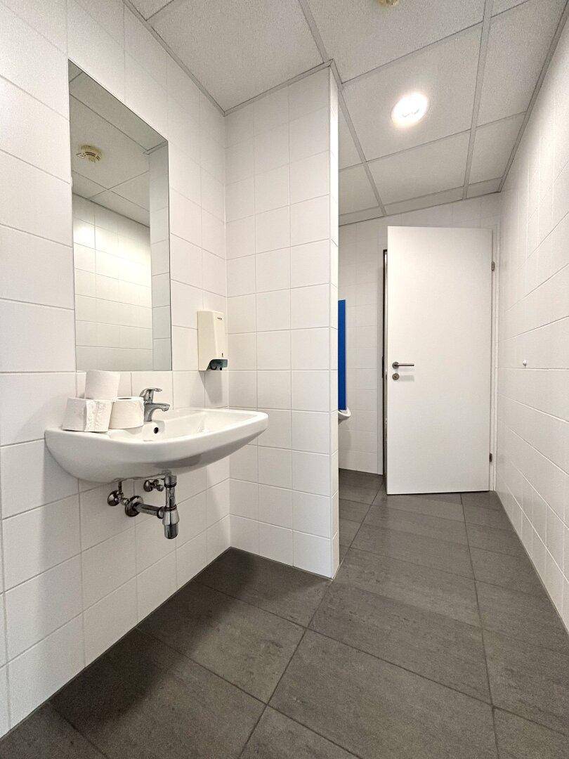 Sauberer Toilettenraum mit weißem Waschbecken, großem Spiegel und hellen Fliesen an den Wänden.