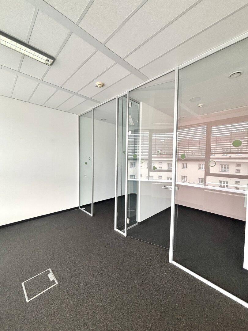 Offener Bürobereich mit Teppichboden und transparenten Glaswänden, die flexible Arbeitsbereiche schaffen.