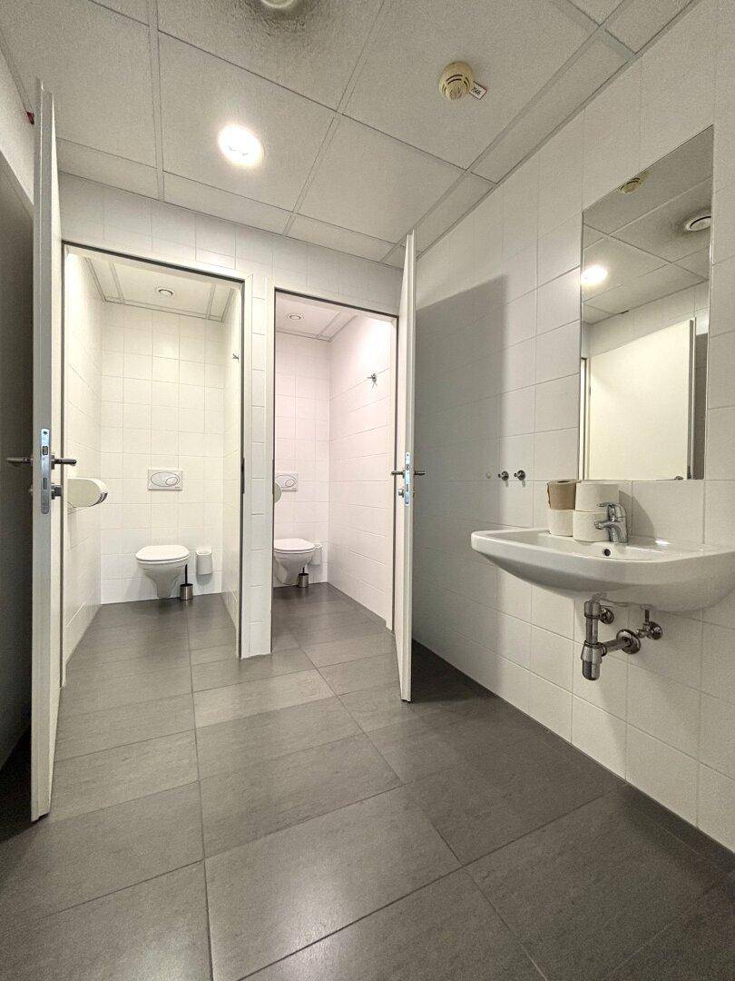 Geräumiger Toilettenraum mit zwei Kabinen, Waschbecken und großem Spiegel, alles hell gefliest.