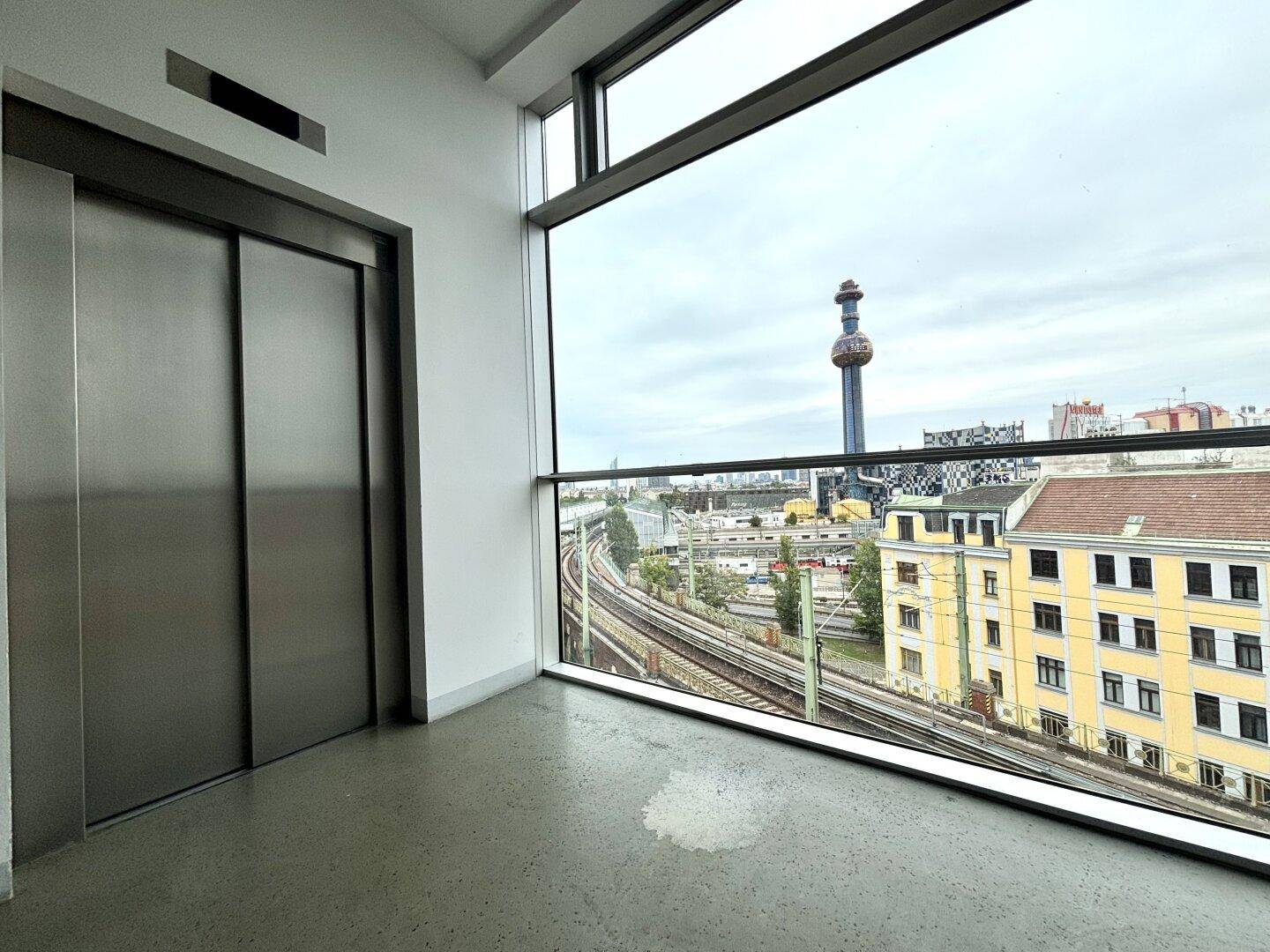 Aufzugsbereich mit Edelstahl-Aufzugtüren und großer Fensterfront mit Blick auf die Stadt und Bahngleise.