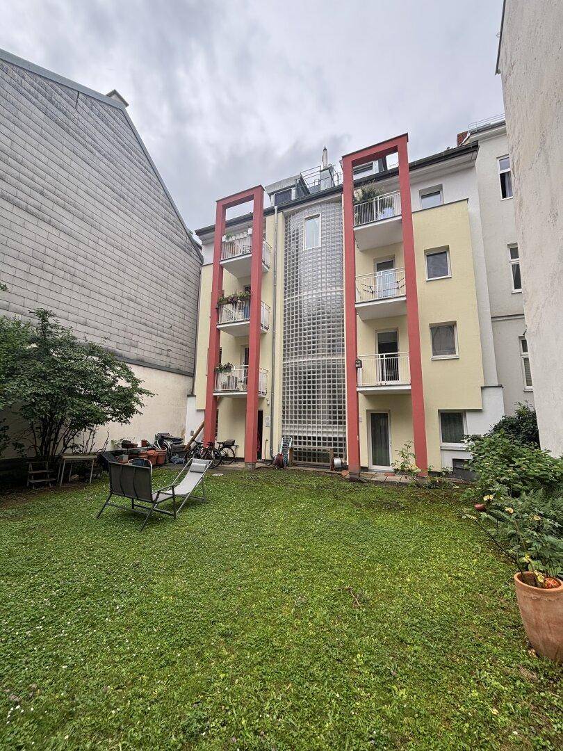 Rückseite des Gebäudes mit Balkonen und Glasbausteinen, umgeben von einem grünen Garten.