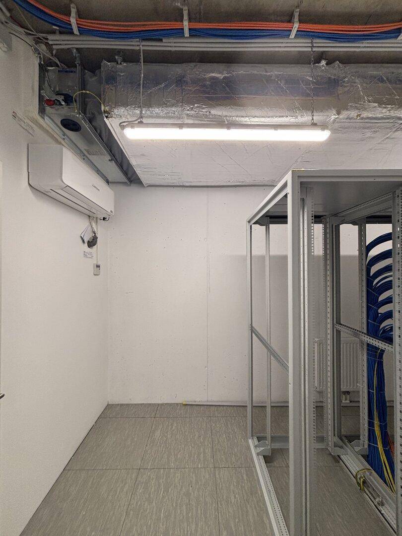 Technischer Raum mit Klimaanlage, Serverschränken und sichtbaren Leitungen an der Decke.