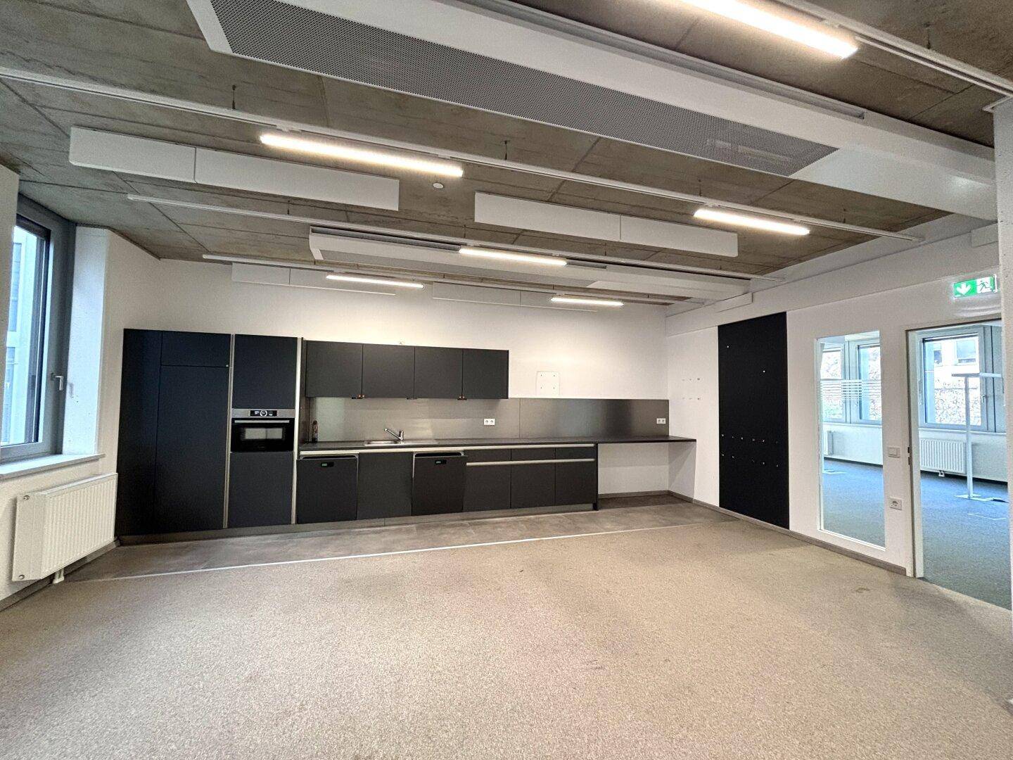 Funktionale Büroküche mit dunklen Schränken, Arbeitsplatte und Zugang zu weiteren Räumen.