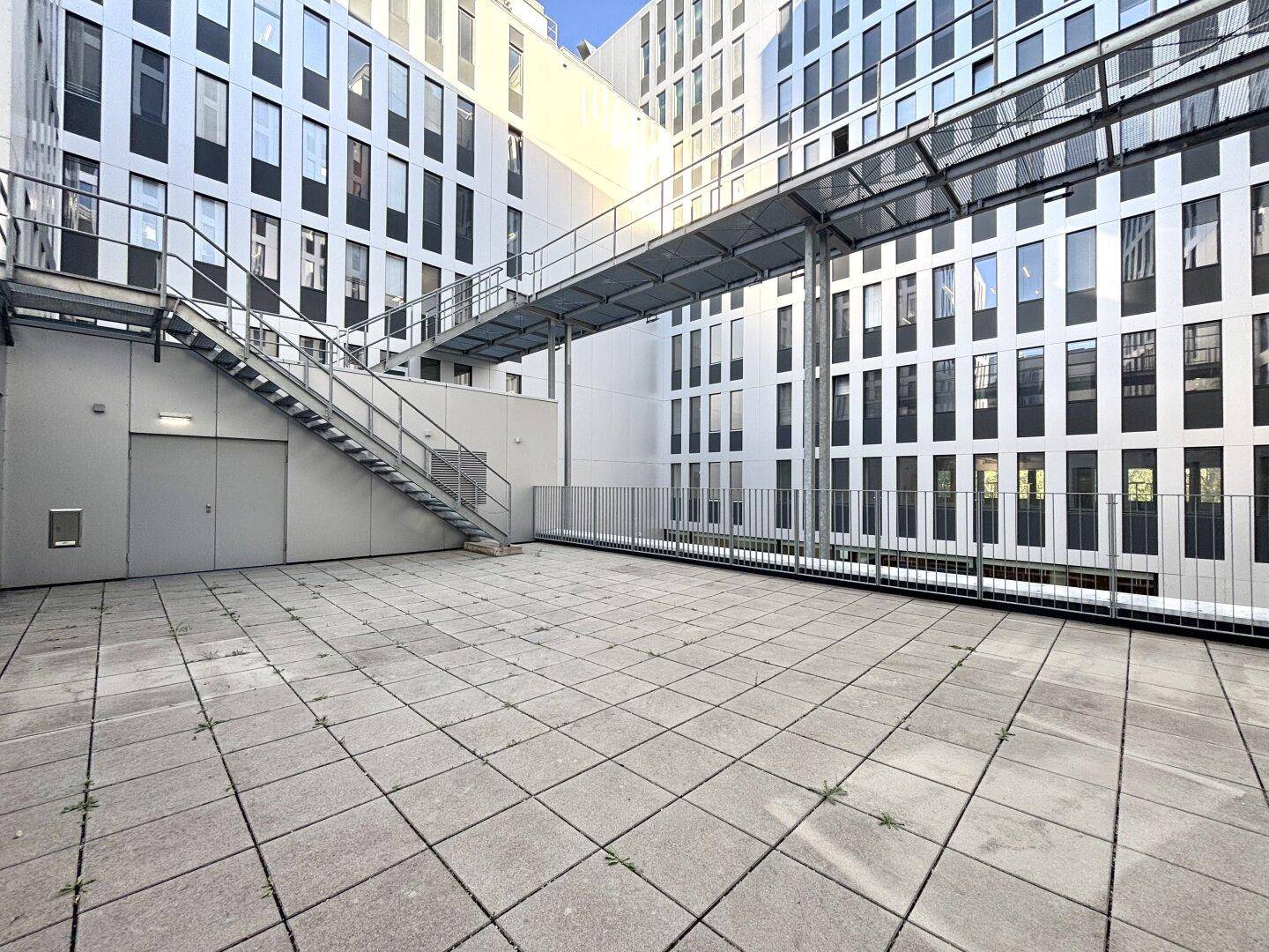 Große gepflasterte Terrasse mit Blick auf moderne Bürogebäude und eine externe Treppe.