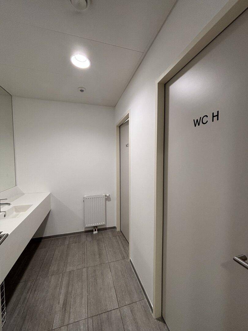 Heller Flur in einem öffentlichen Waschraum mit grauen Fliesen und einer Tür zum WC.