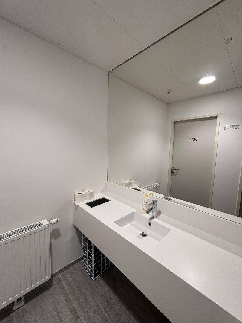 Sauberer Waschraum mit langem, weißem Waschtisch, großem Spiegel und Heizkörper unter dem Fenster.
