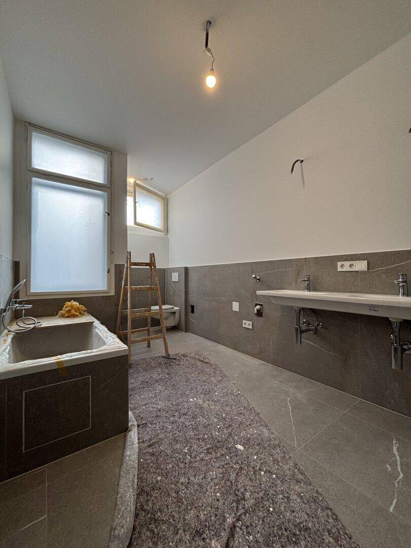 Geräumiges Badezimmer in Renovierung mit Badewanne, Doppelwaschbecken und modernen grauen Fliesen.
