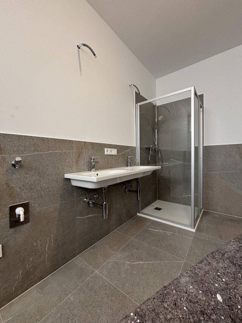 Modernes Badezimmer in Renovierung mit Duschkabine, Doppelwaschbecken und grauen Wand- und Bodenfliesen.