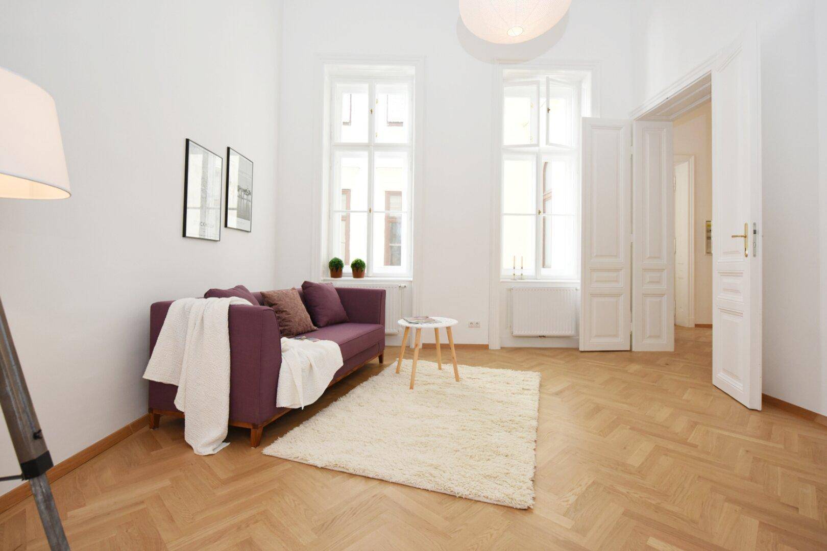 Gemütliches Wohnzimmer mit Fischgrätparkett, lila Sofa und hohen Fenstern.