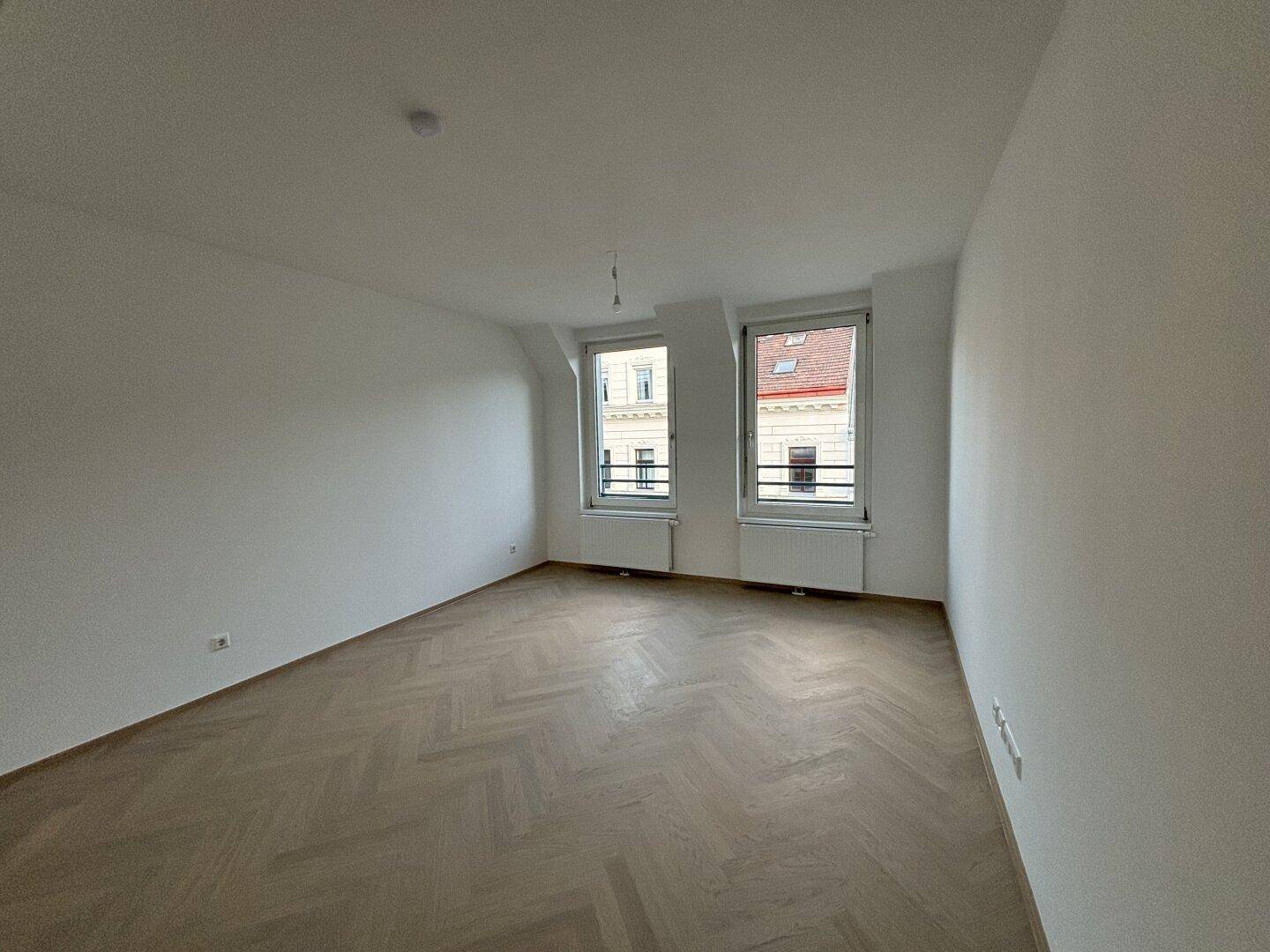 Helles, unmöbliertes Schlafzimmer mit Fischgrätparkett und zwei Fenstern.