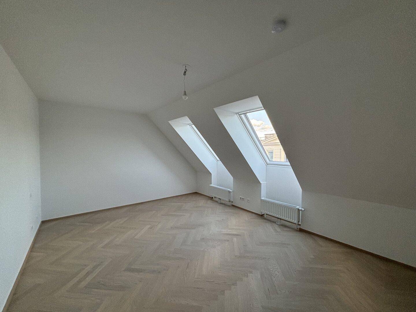 Geräumiges Dachgeschosszimmer mit zwei Dachfenstern, schrägen Wänden und Fischgrätparkett.