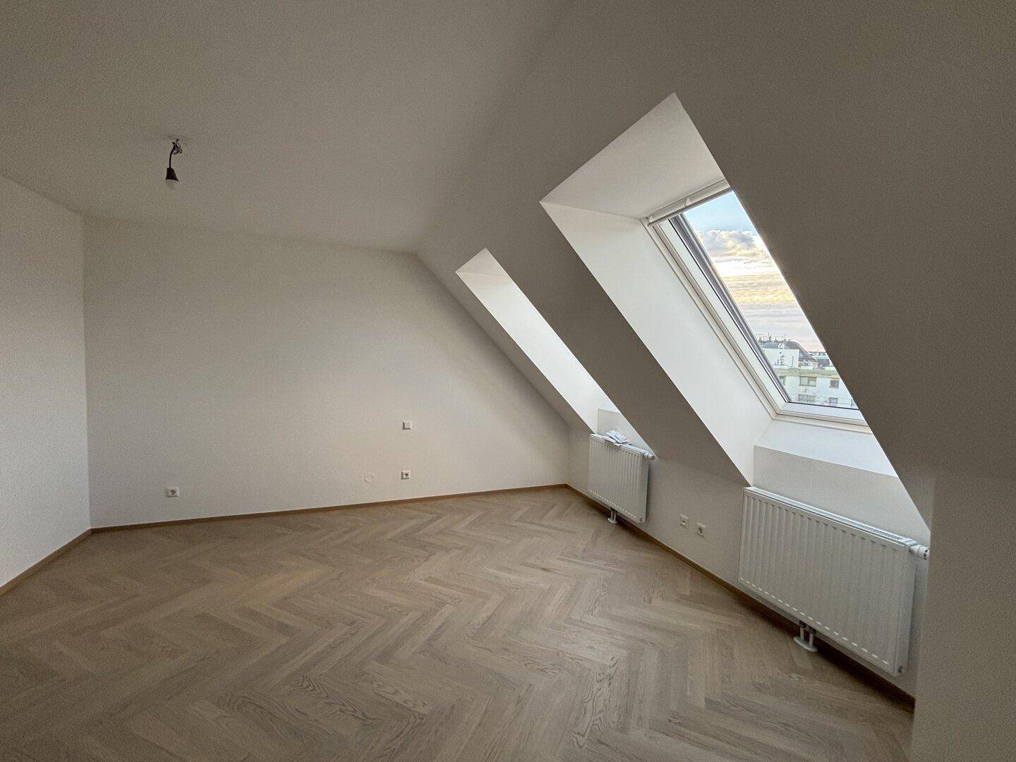 Helles Dachgeschosszimmer mit schrägen Wänden, zwei Dachfenstern und Fischgrätparkett.