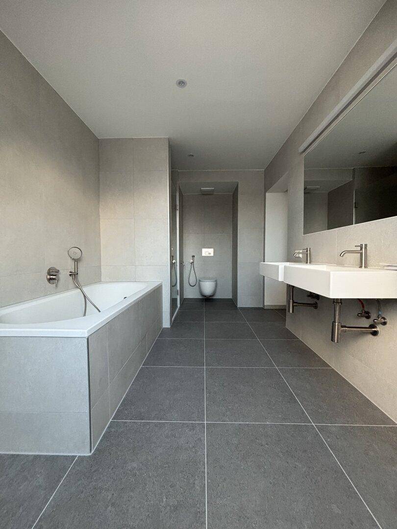 Helles Badezimmer mit Doppelwaschbecken, Badewanne, Toilette und modernen grauen Fliesen.