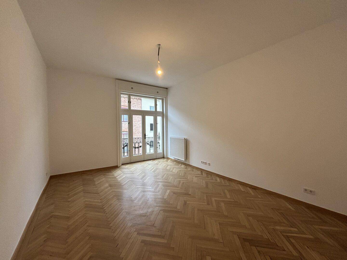 Heller, unmöblierter Raum mit klassischem Fischgrätparkett und Balkonzugang, ideal für ein Wohnzimmer.