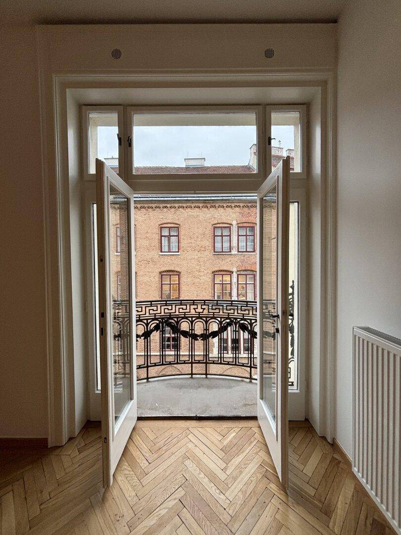 Blick durch die geöffneten Balkontüren auf einen kleinen Balkon mit schmiedeeisernem Geländer und gegenüberliegendem Gebäude.