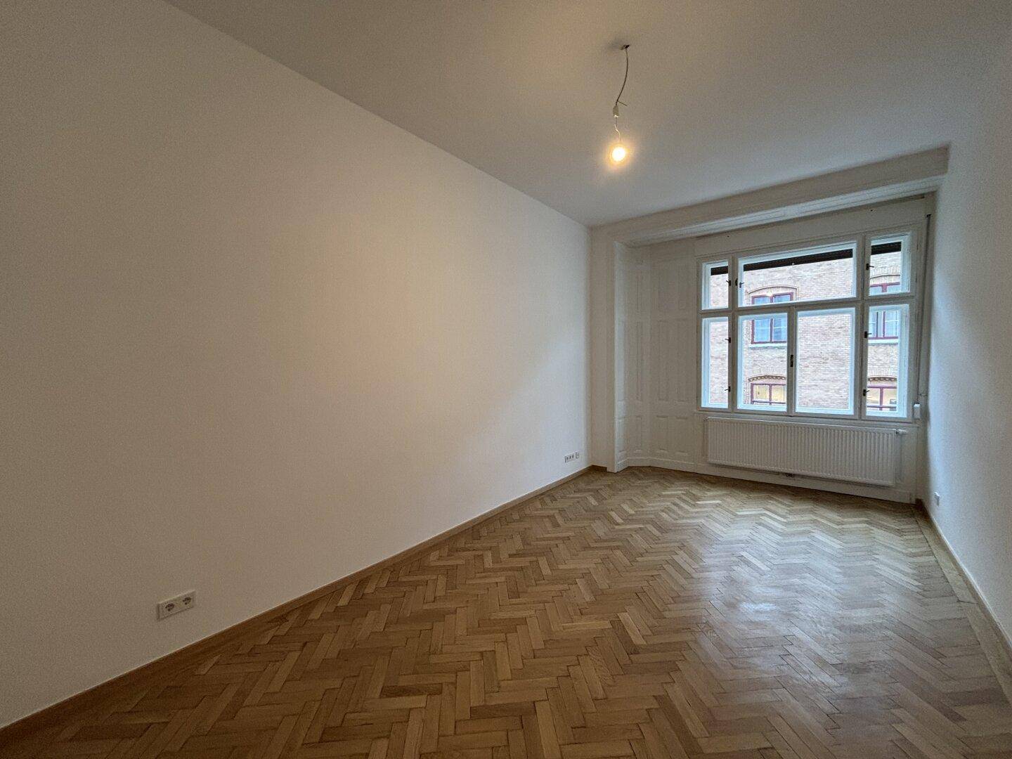 Geräumiger Raum mit Fischgrätparkett und großem Fenster, ideal als Schlafzimmer oder Arbeitszimmer.