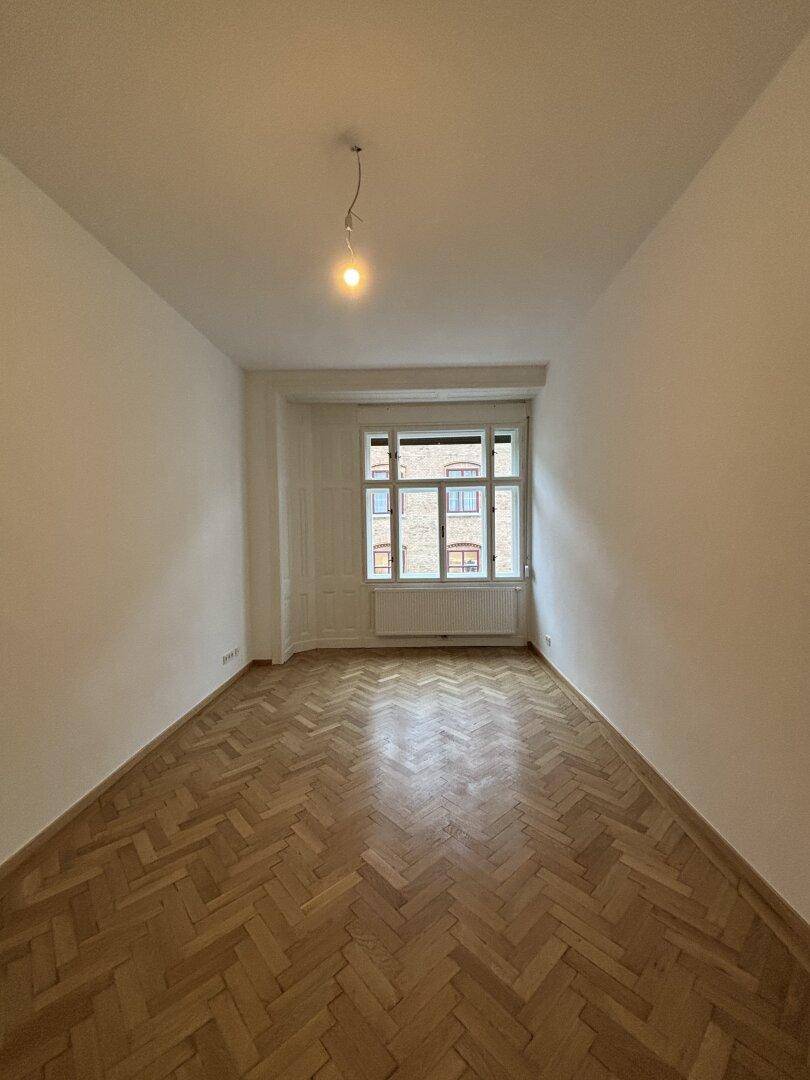 Weitläufiger, unmöblierter Raum mit Fischgrätparkett und großem Fenster, ideal als Schlafzimmer.