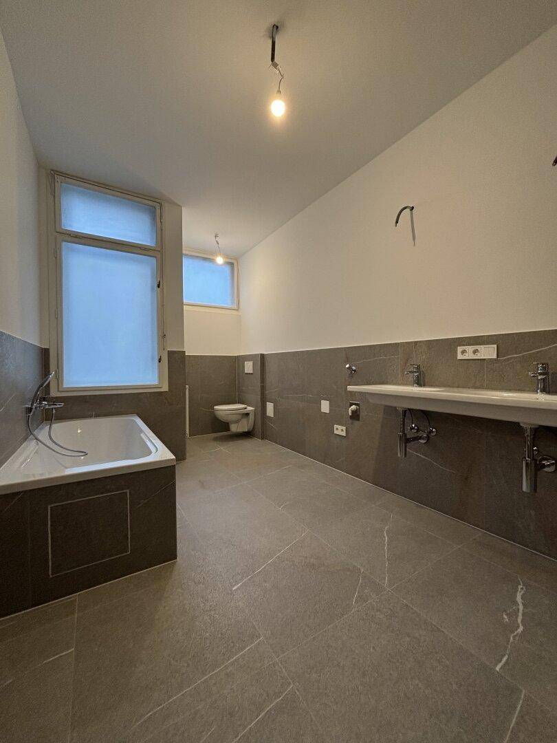 Modernes Badezimmer mit grauen Fliesen, Badewanne, wandhängendem WC und Doppelwaschbecken.